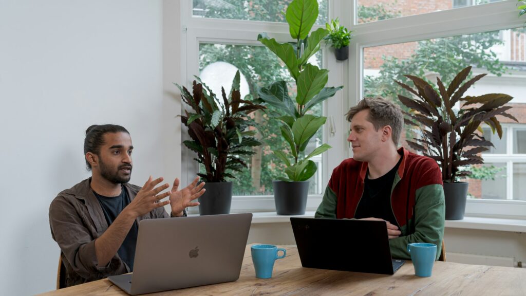 Two colleagues talking across a table with laptops in a bright office